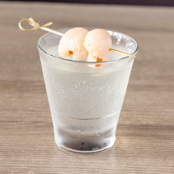 A clear glass of cold beverage with two lychee fruits on a skewer as garnish.