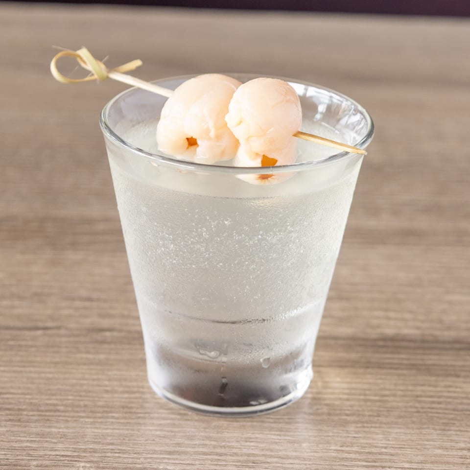 A clear glass of cold beverage with two lychee fruits on a skewer as garnish.