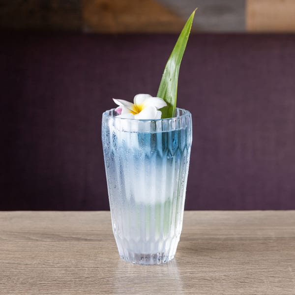 A refreshing blue cocktail in a textured glass, garnished with a white flower and green leaf.