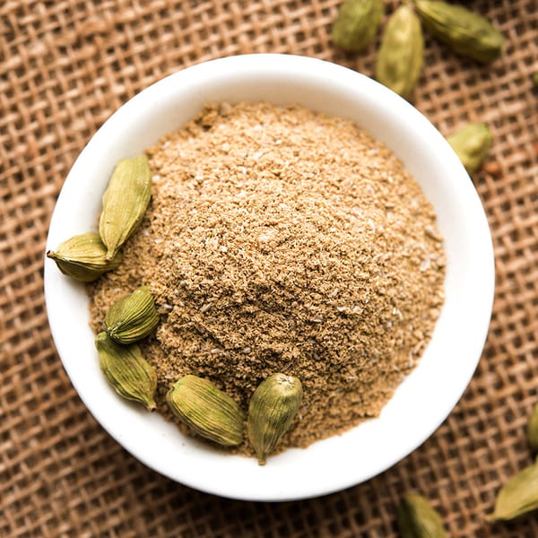 A white bowl filled with ground cardamom powder, surrounded by whole green cardamom pods on burlap.